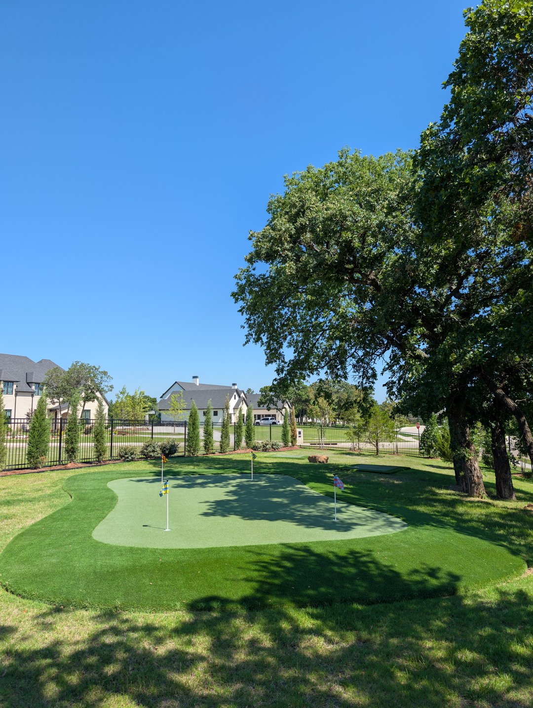 Backyard putting green with four flags beside a fenced yard and shade trees