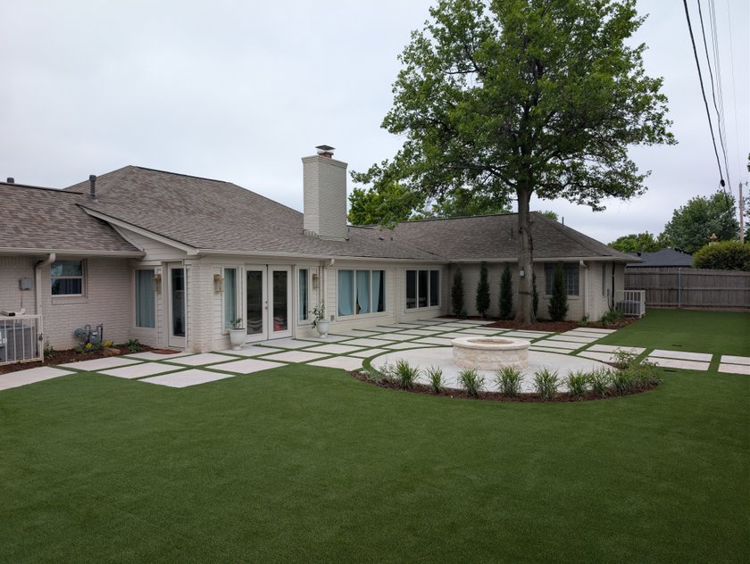 Artificial turf lawn around a paver patio with turf strips and a round stone fire pit behind a white brick home