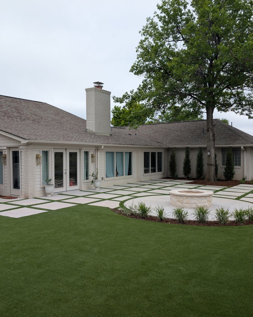 Artificial turf lawn around a paver patio with turf strips and a round stone fire pit behind a white brick home