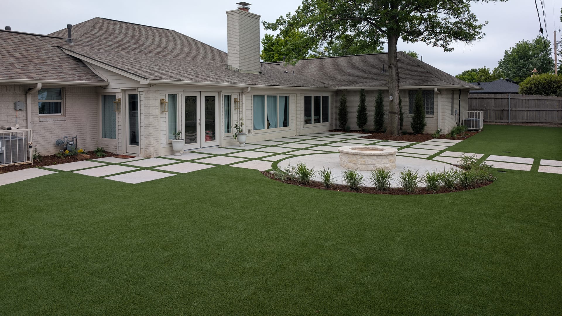 Artificial turf lawn around a paver patio with turf strips and a round stone fire pit behind a white brick home