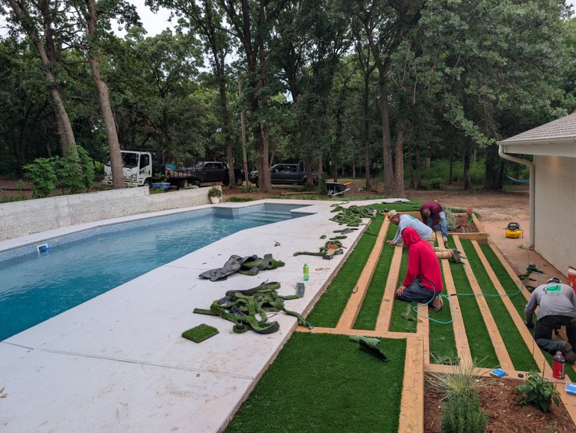 Crew cutting artificial turf strips between edging boards beside a new pool deck