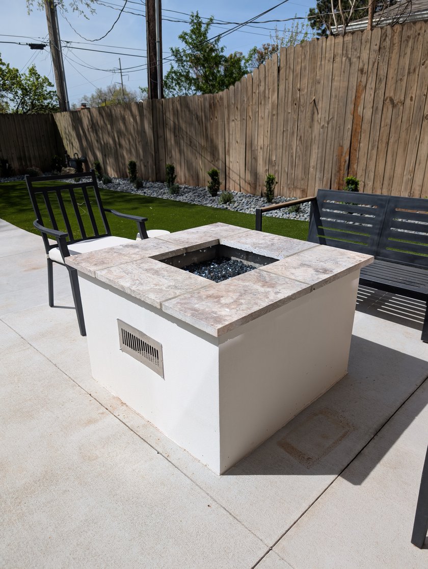 Square stucco fire pit table on a concrete patio next to artificial turf and a cedar fence