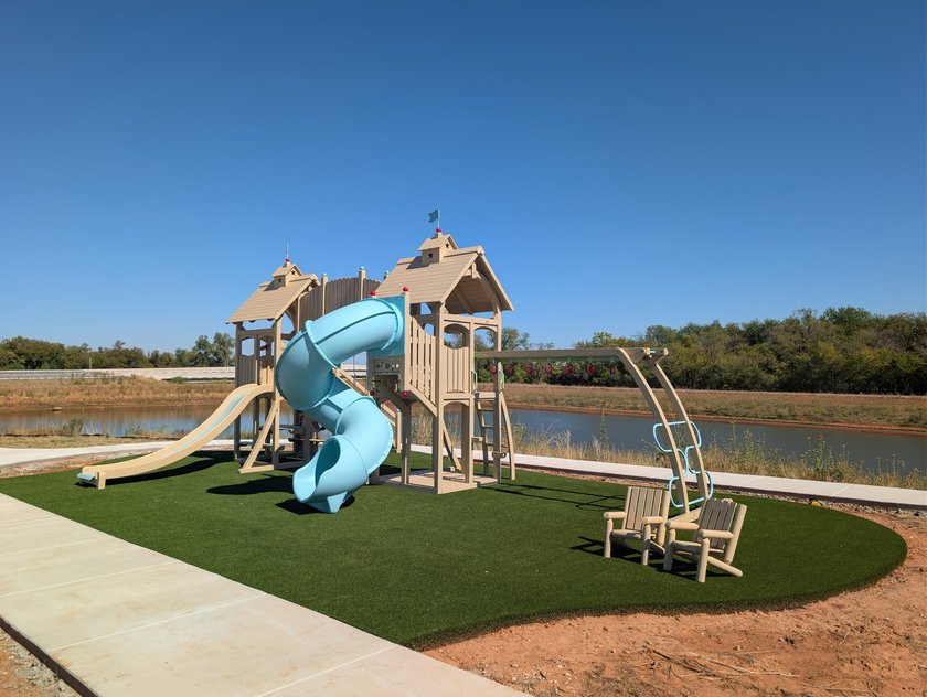 Artificial turf playground pad with a slide tower, tire swings and two chairs, edged by a concrete walk along a pond