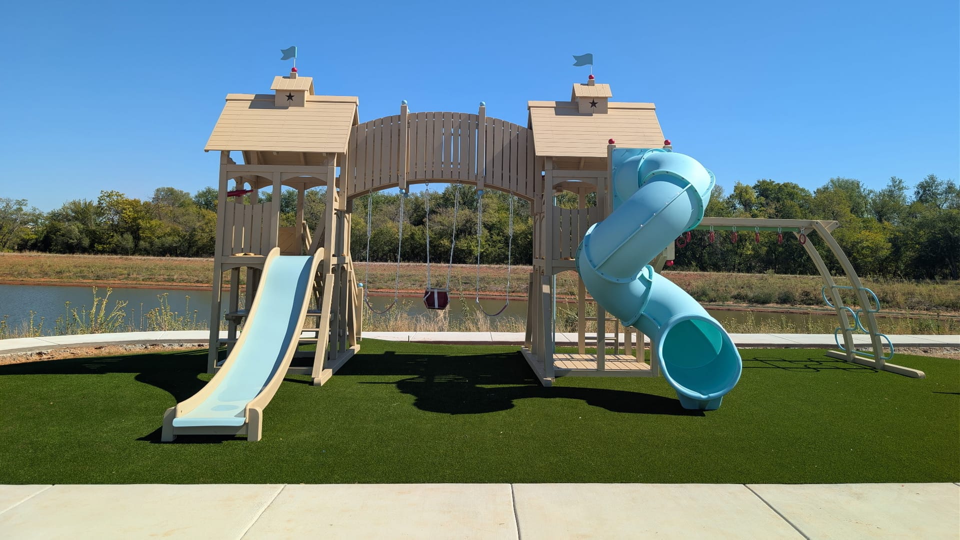 Playground with two towers, a spiral slide and swings set on an artificial turf pad beside a pond