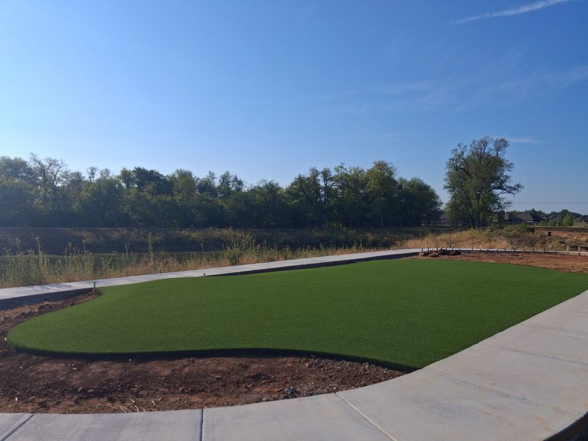 Artificial turf lawn curving between concrete walks, with a pond and treeline behind it