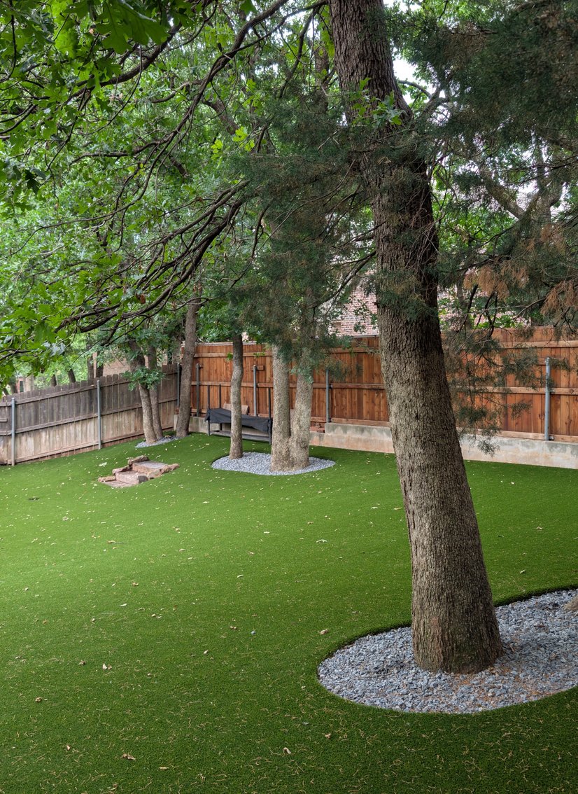 Pet turf installed around mature shade trees in a fenced backyard, with river rock rings around each trunk