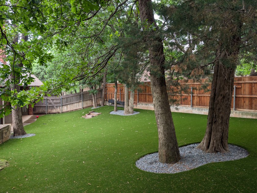 Pet turf installed around mature shade trees in a fenced backyard, with river rock rings around each trunk