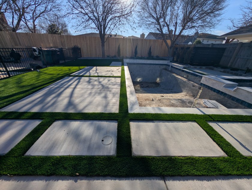 Artificial turf strips installed between large concrete pavers beside a pool under construction