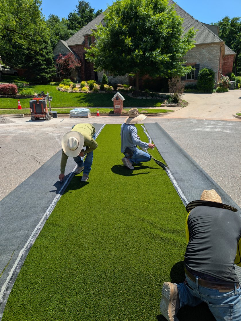 Crew kneeling to lay and trim a long strip of artificial turf beside a driveway